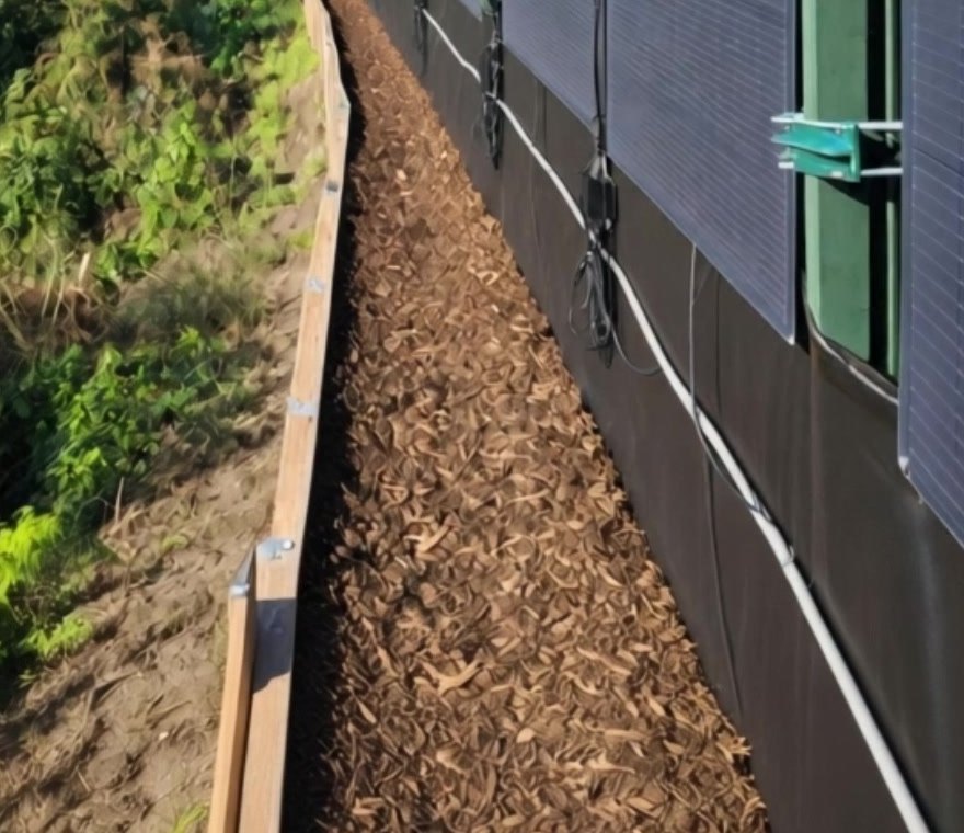 Paneles solares flexibles sujetos con soportes a postes de valla de acero verde sobre un camino de astillas de madera